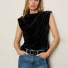 A woman with long blonde hair wears the Sanctuary Clothing pleated sleeveless top in black and blue jeans, accessorized with a black belt, standing against a neutral background with a relaxed expression.