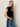 A woman with long blonde hair models the Sanctuary Clothing pleated sleeveless top in black and blue side-striped jeans, standing against a light background with her hands in her pockets, looking at the camera.