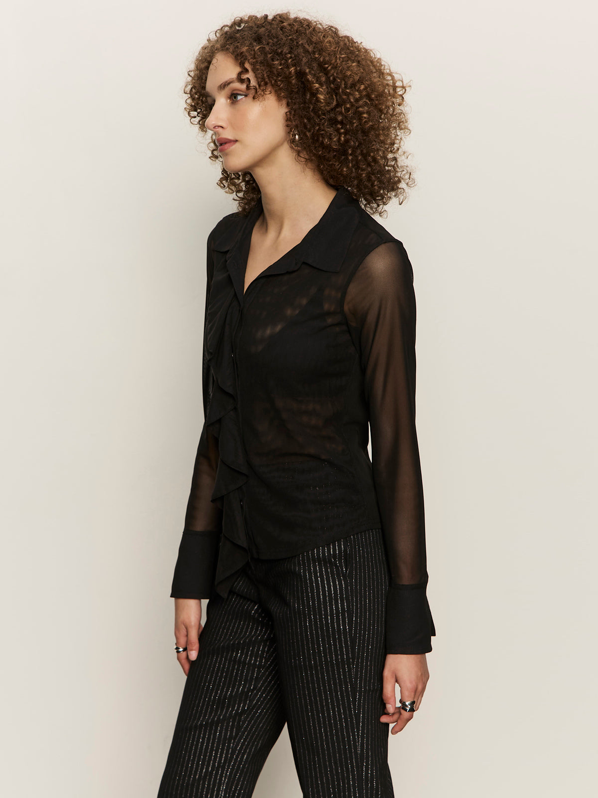 A person with curly hair models the Sanctuary Clothing dreamland ruffle top black, paired with black pinstriped pants, standing against a plain light background and looking to the side.