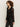 A person with curly hair models the Sanctuary Clothing dreamland ruffle top black, paired with black pinstriped pants, standing against a plain light background and looking to the side.