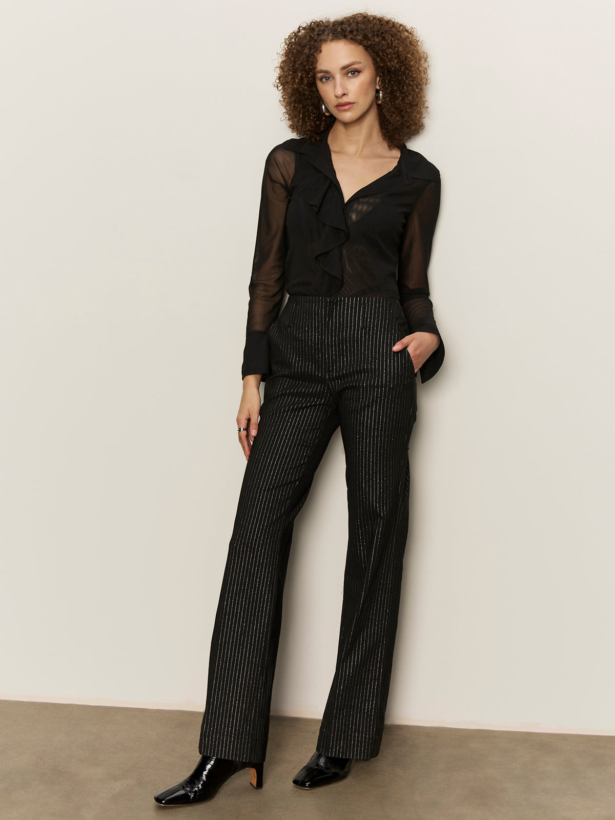 A woman with curly hair models the Sanctuary Clothing dreamland ruffle top in black, paired with high-waisted pinstriped pants and black pointed-toe shoes, standing against a plain wall with one hand in her pocket.