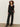 A woman with curly hair models the Sanctuary Clothing dreamland ruffle top in black, paired with high-waisted pinstriped pants and black pointed-toe shoes, standing against a plain wall with one hand in her pocket.