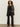A person stands against a beige wall wearing Sanctuary Clothing„¢s dreamland ruffle top in black, paired with a leopard print coat, black pinstriped trousers, and black heeled boots. They have curly hair and a neutral expression.