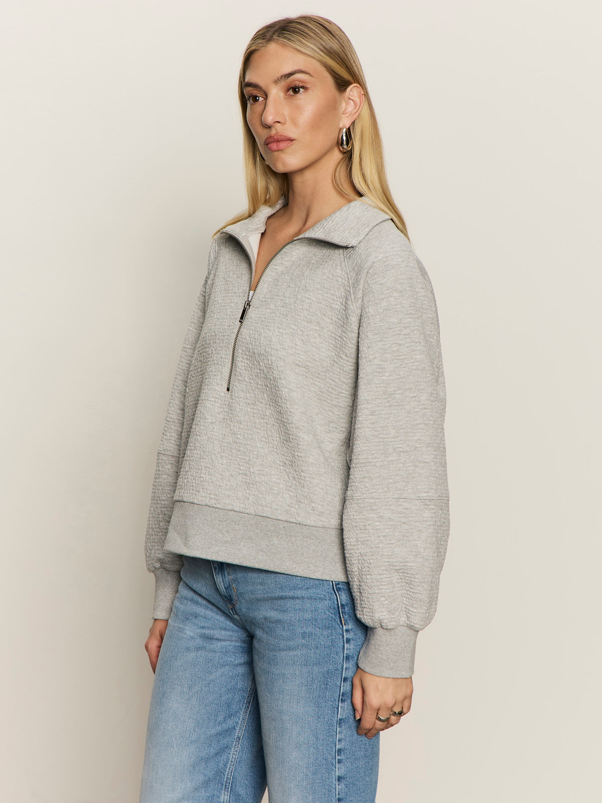 A woman with long blonde hair wears the Sanctuary Clothing textured zip up popover in heather grey and blue jeans, standing against an off-white background and looking slightly to the side.