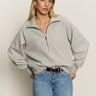 A woman with long blonde hair wears the Sanctuary Clothing textured zip up popover in heather grey and blue jeans, standing with one hand in her pocket against a plain, light background.