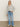 A woman with straight blonde hair and a neutral expression stands against a light wall, wearing Sanctuary Clothing„¢s textured zip up popover in heather grey, paired with light blue jeans, white sneakers, hoop earrings, and one hand in her pocket.
