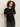 A woman with curly brown hair wears the Sanctuary Clothing satin mix tee in black, paired with black jeans, as she stands against an off-white background and gazes slightly to the side with a neutral expression.