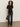 A woman with curly hair wears the Sanctuary Clothing satin mix tee in black, paired with straight-leg dark jeans and pointed black shoes. She holds a small black handbag while standing against a plain beige background.