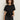 A woman with curly hair wears the Sanctuary Clothing satin mix tee in black, featuring gathered detailing on one side, paired with dark jeans. She stands against a plain light background, looking at the camera with a neutral expression.