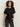 A woman with curly hair wears the Sanctuary Clothing satin mix tee in black, featuring gathered detailing on one side, paired with dark jeans. She stands against a plain light background, looking at the camera with a neutral expression.