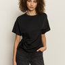 A woman with curly hair wears the Sanctuary Clothing satin mix tee in black, featuring gathered detailing on one side, paired with dark jeans. She stands against a plain light background, looking at the camera with a neutral expression.