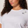 A woman with curly brown hair wears the Sanctuary Clothing embellished tee in white (extended sizing) featuring sparkling jeweled details, paired with gold hoop earrings, standing against a neutral background with a calm expression.