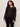 A woman with curly hair models Sanctuary Clothing’s lovely lace button front top in black (extended sizing) with black pants, standing against a light background and looking confidently at the camera.