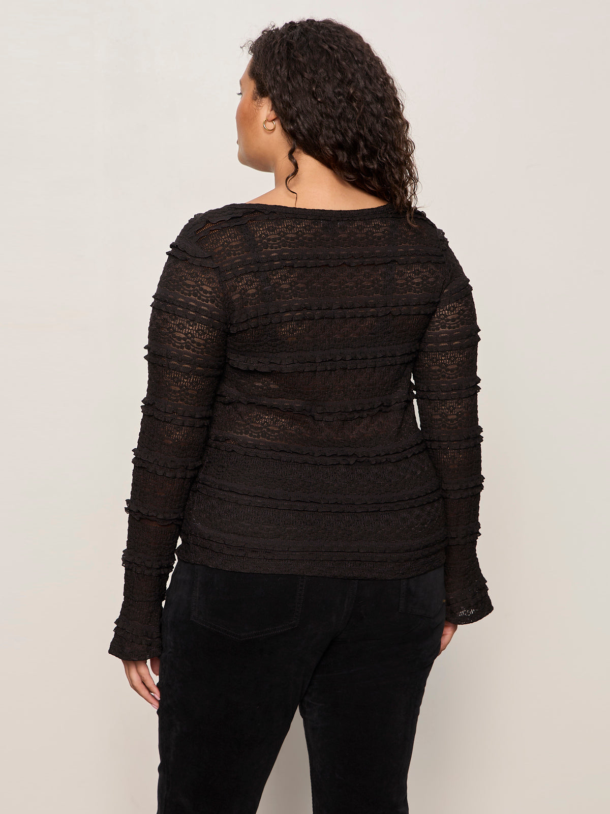 A woman with curly hair faces away, wearing Sanctuary Clothing's lovely lace button front top in black extended sizing with black pants, set against a plain light background.