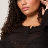 A woman with curly brown hair and gold hoop earrings models the Sanctuary Clothing lovely lace button front top in black extended sizing, featuring sheer horizontal stripes. She poses confidently, raising one hand to her ear.