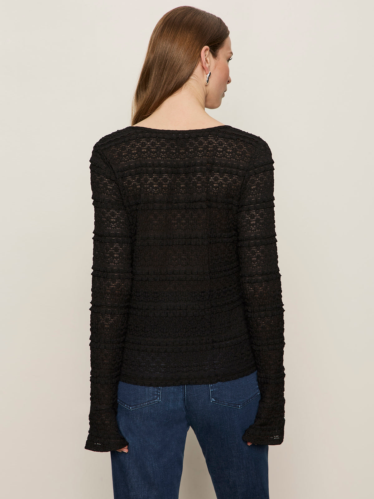 A woman with long brown hair is seen from the back, wearing the Sanctuary Clothing lovely lace button front top in black and blue jeans, standing against a plain light background.