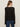 A woman with long brown hair is seen from the back, wearing the Sanctuary Clothing lovely lace button front top in black and blue jeans, standing against a plain light background.