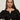 A woman with long brown hair wears the Sanctuary Clothing lovely lace button front top in black and silver hoop earrings, sitting on a light-colored chair. She looks at the camera with a neutral expression, her hand showing silver rings.