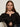 A woman with long brown hair wears the Sanctuary Clothing lovely lace button front top in black and silver hoop earrings, sitting on a light-colored chair. She looks at the camera with a neutral expression, her hand showing silver rings.