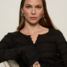 A woman with long brown hair wears the Sanctuary Clothing lovely lace button front top in black and silver hoop earrings, sitting on a light-colored chair. She looks at the camera with a neutral expression, her hand showing silver rings.