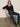 A woman with long brown hair sits on a cream chair, wearing the Sanctuary Clothing lovely lace button front top in black, paired with blue jeans, a black belt, and black boots. She poses against a plain background, looking directly at the camera.