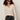 A woman with curly hair wears black sunglasses, black jeans, and a black belt. She stands against a plain light background in the Sanctuary Clothing lovely lace button front top in vintage ivory.