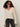 A woman with curly hair wears black sunglasses, black jeans, and a black belt. She stands against a plain light background in the Sanctuary Clothing lovely lace button front top in vintage ivory.