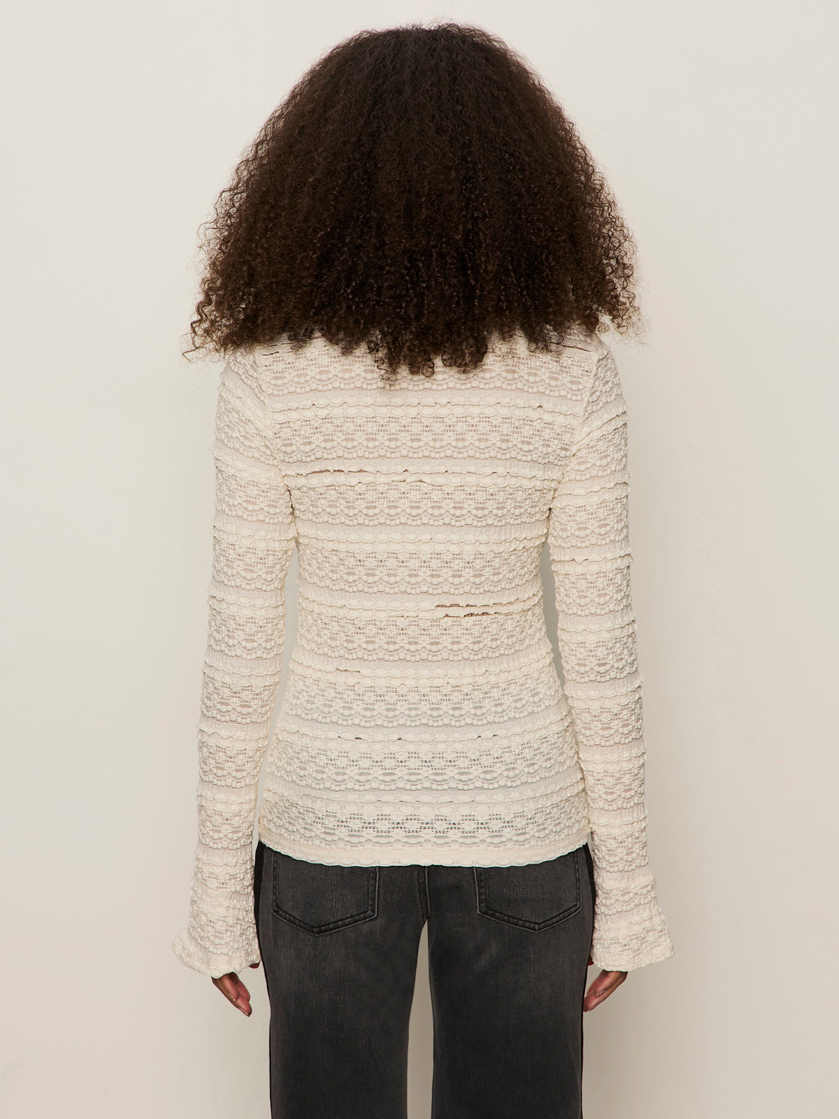 A person with curly hair is shown from the back, wearing the Sanctuary Clothing lovely lace button front top in vintage ivory and dark jeans, standing against a plain light background.