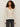 A woman with curly hair wears the Sanctuary Clothing lovely lace button front top in vintage ivory and black jeans, standing against a plain light background.