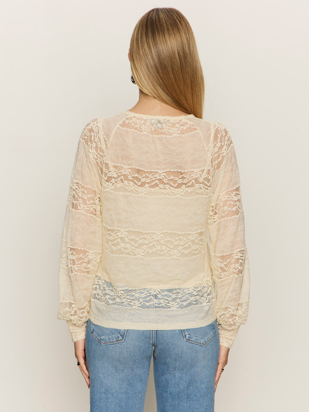 A woman with straight blonde hair models the Sanctuary Clothing ultra femme raglan top in vintage ivory, featuring sheer lace long sleeves, paired with light blue jeans. She stands with her back to the camera against a plain light background.