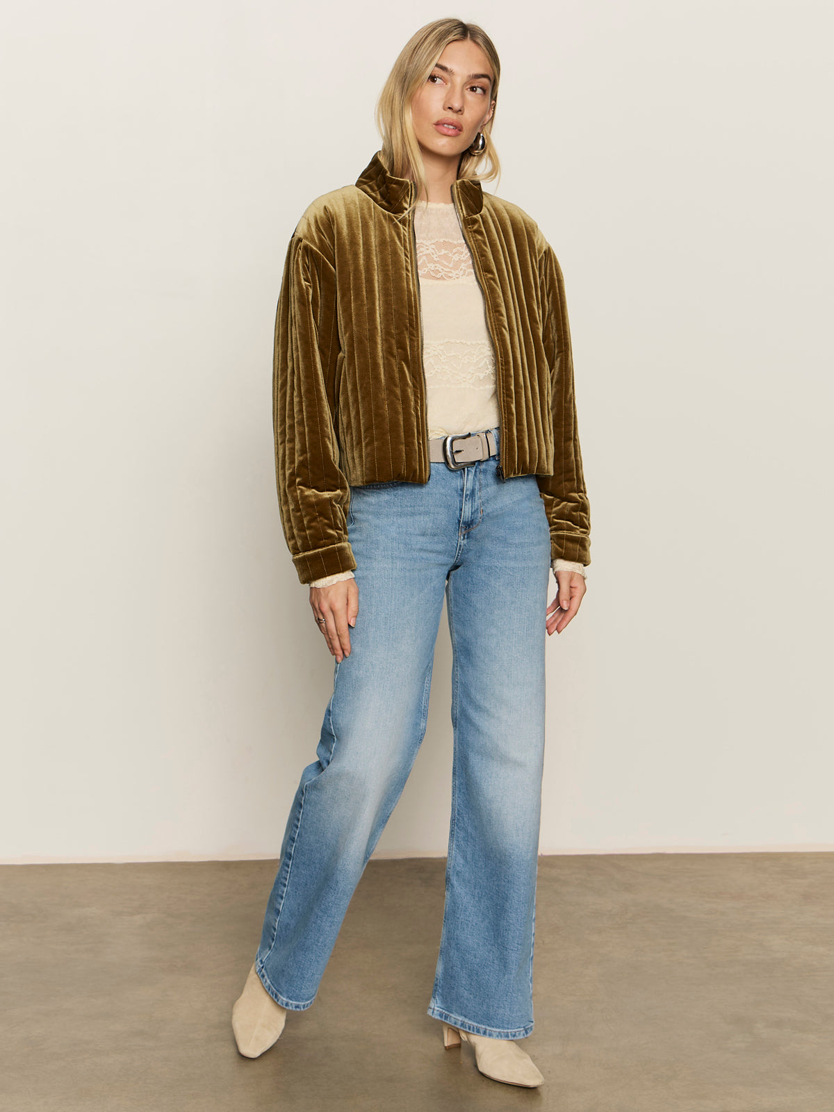 A woman with blonde hair stands against a neutral background wearing Sanctuary Clothing„¢s ultra femme raglan top in vintage ivory, styled with light blue high-waisted jeans, a beige belt, tan velvet jacket, and beige pointed-toe boots.