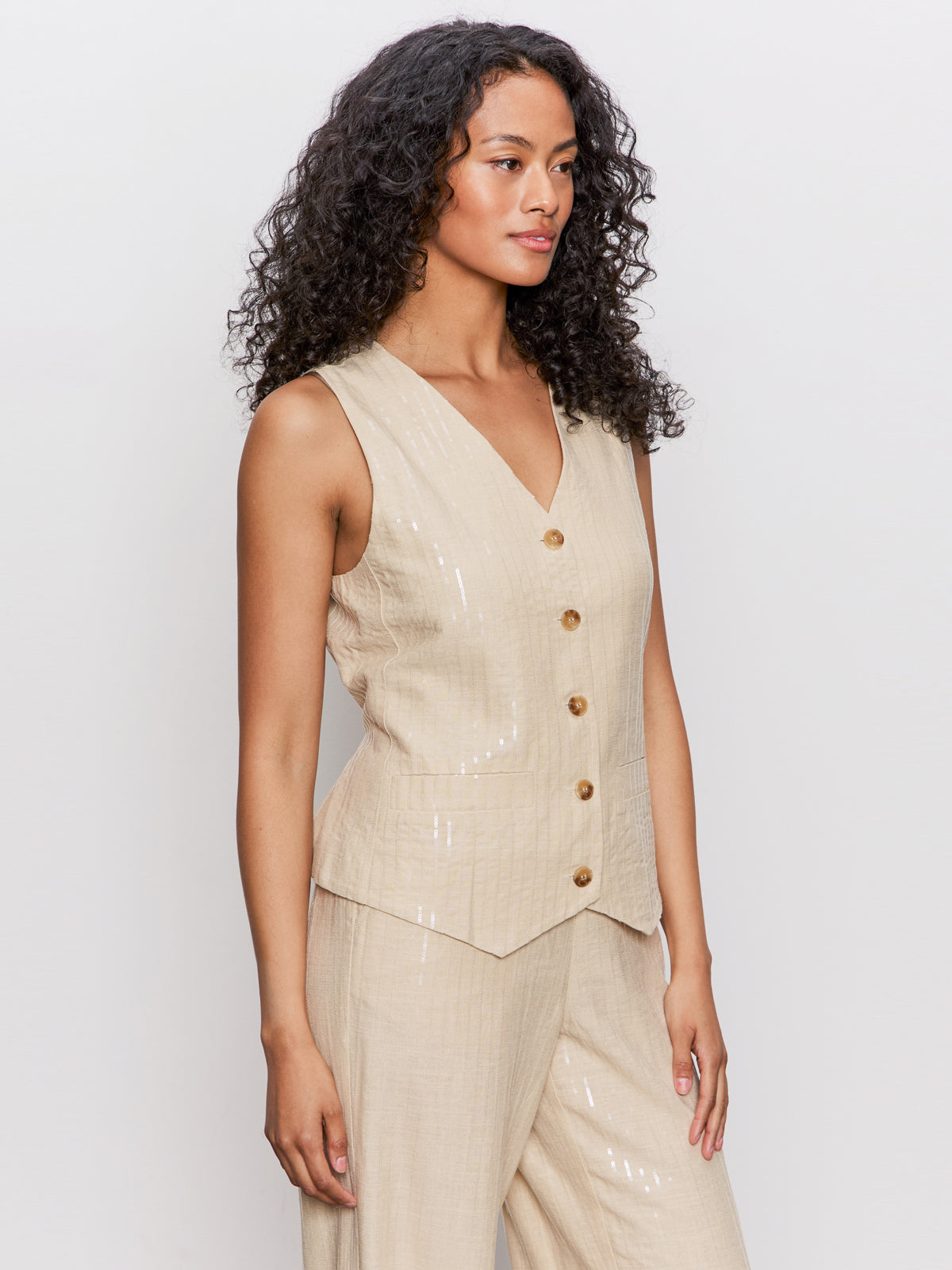 A woman with long curly hair wears the Sanctuary Clothing relaxed modern vest sand sequin stripe and matching wide-leg pants, standing against a plain light background and looking slightly to the side.