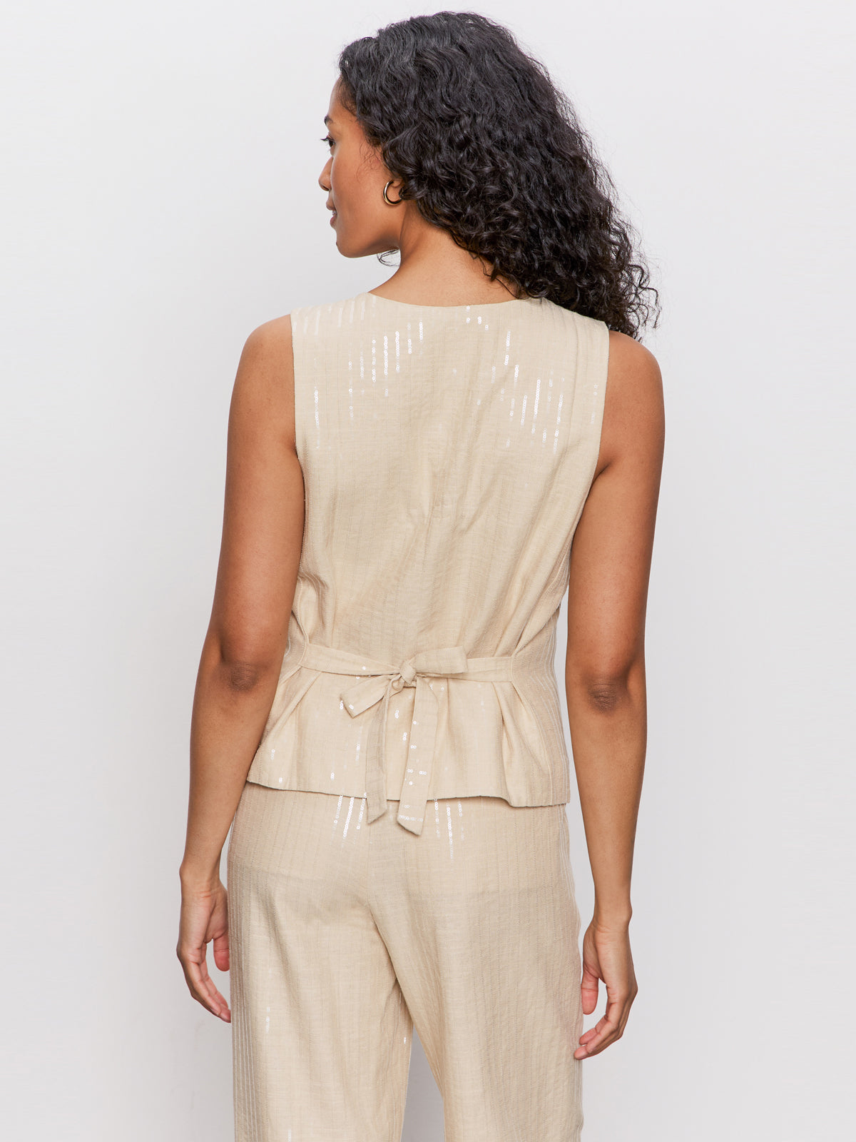 A woman with long curly hair faces away from the camera, wearing the Sanctuary Clothing relaxed modern vest in sand sequin stripe—a sleeveless beige top with a tied waist and subtle shiny pattern, styled with matching beige pants.