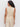 A woman with long curly hair faces away from the camera, wearing the Sanctuary Clothing relaxed modern vest in sand sequin stripe�a sleeveless beige top with a tied waist and subtle shiny pattern, styled with matching beige pants.