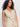 A woman with long curly hair models the Sanctuary Clothing relaxed modern vest sand sequin stripe, a beige sleeveless button-up vest with a subtle sheen, standing against a plain white background.