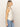 A person with long blonde hair stands sideways in a sleeveless, fitted relaxed modern vest natural metallic stripe by Sanctuary Clothing paired with light blue jeans against a plain, light background.