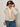 A woman wearing dark sunglasses, blue jeans, and a white long-sleeve shirt stands against a plain light background. She holds the collar of her Sanctuary Clothing silky fur vest almond cream (extended sizing).