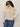 A woman in Sanctuary Clothing's silky fur vest in almond cream (extended sizing) over a ribbed white long-sleeve top, blue jeans, and dark sunglasses stands against a plain light background.