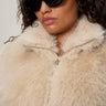 A woman with curly hair wears black oval sunglasses and the Sanctuary Clothing silky fur vest in almond cream (extended sizing), featuring a zippered collar.