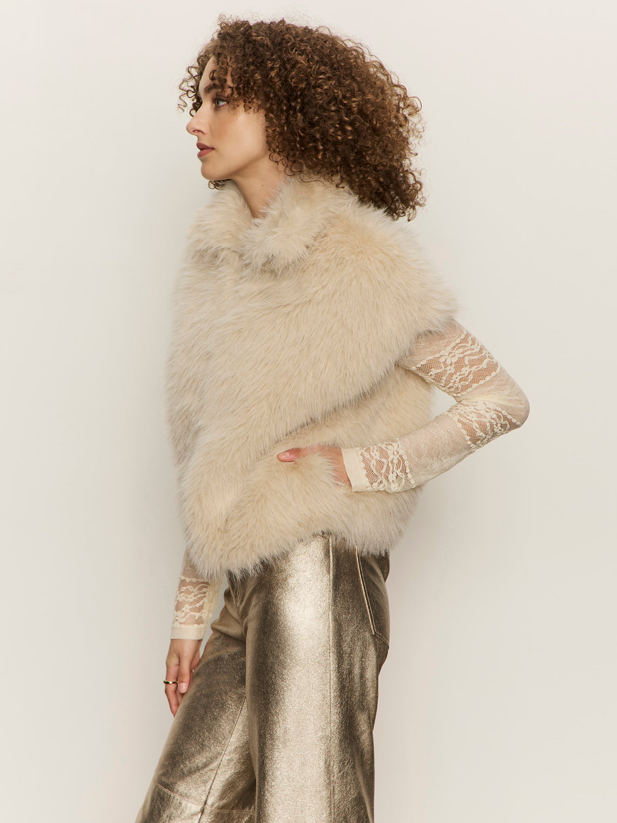 A person with curly hair models the silky fur vest in almond cream by Sanctuary Clothing over a sheer lace top and metallic gold pants, standing in profile against a plain light background.