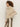 A person with curly hair models the silky fur vest in almond cream by Sanctuary Clothing over a sheer lace top and metallic gold pants, standing in profile against a plain light background.