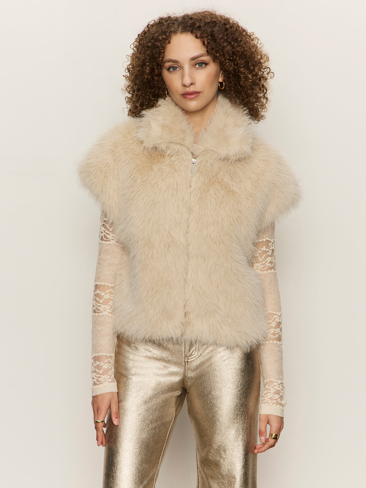 A woman with curly hair wears the Sanctuary Clothing silky fur vest in almond cream over a sheer lace long-sleeve top and metallic gold pants, standing against a plain light background.