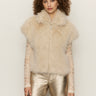 A woman with curly hair wears the Sanctuary Clothing silky fur vest in almond cream (petite sizing) over a sheer, long-sleeved lace top and metallic gold pants, standing against a plain light background.