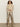 Someone with curly hair wears the Sanctuary Clothing silky fur vest in almond cream over a sheer long-sleeve top, paired with gold metallic flared pants and beige heels, standing against a plain off-white wall.