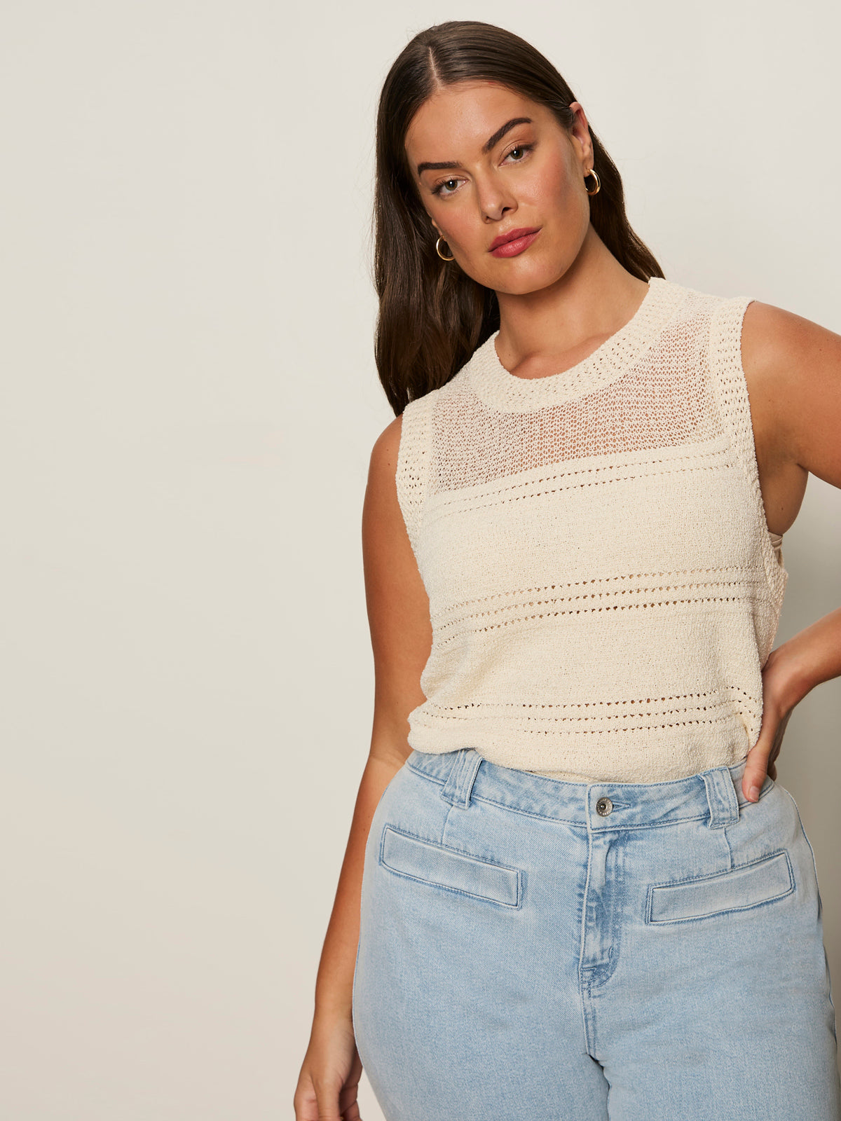 A woman with long brown hair wears the Sanctuary Clothing "waves of summer shell sweater macrame" in extended sizing, paired with light blue high-waisted jeans, standing against a plain light background with a neutral expression.