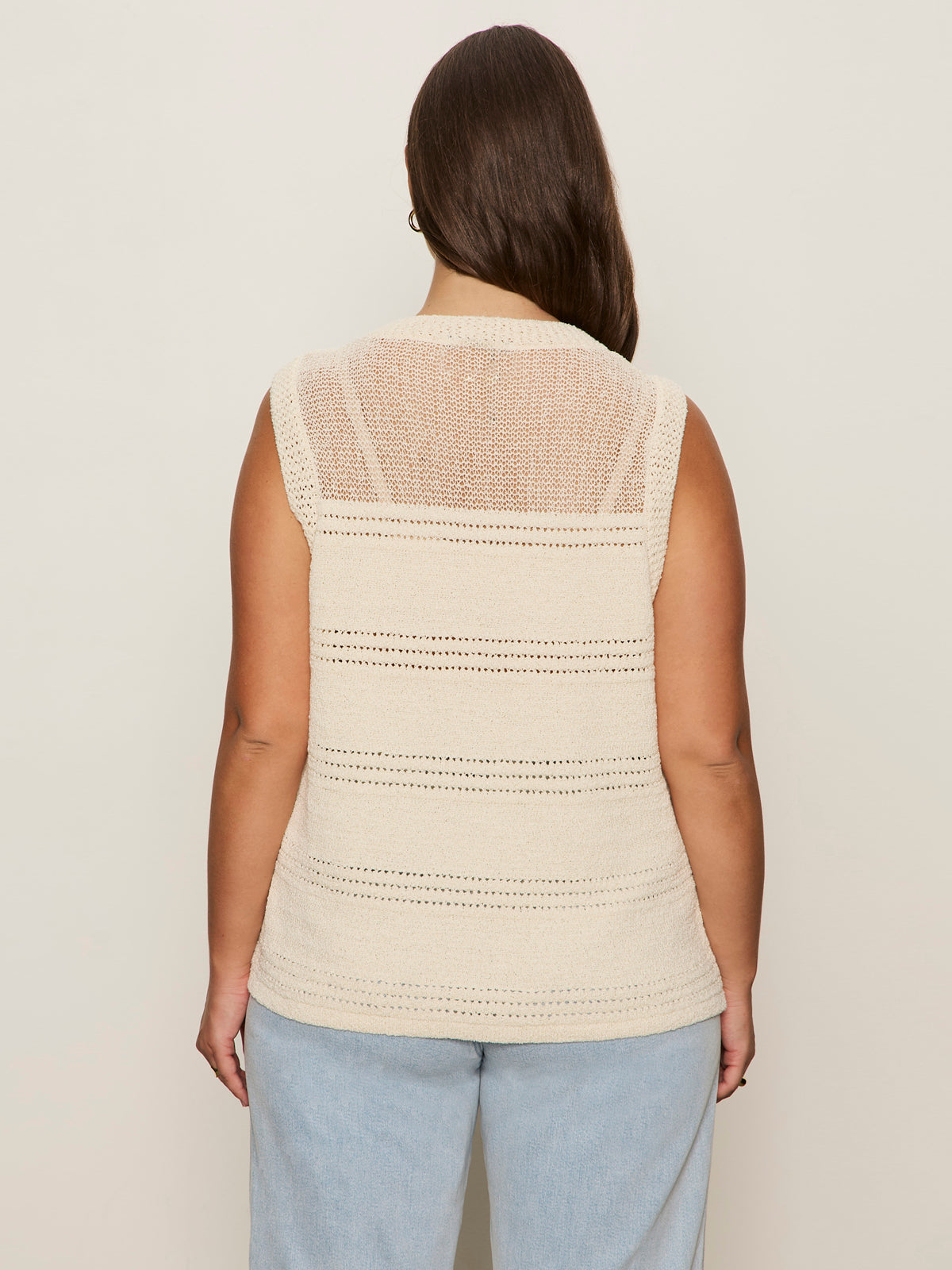 A person with long brown hair is shown from the back against a beige background, wearing Sanctuary Clothing's waves of summer shell sweater macrame in extended sizing—a sleeveless, cream knit top—paired with light blue jeans.