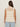 A person with long brown hair is shown from the back against a beige background, wearing Sanctuary Clothing's waves of summer shell sweater macrame in extended sizing—a sleeveless, cream knit top—paired with light blue jeans.