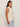 A woman with long brown hair models the Sanctuary Clothing "waves of summer shell sweater macrame" in extended sizing, paired with light blue jeans, standing sideways against a plain beige background.