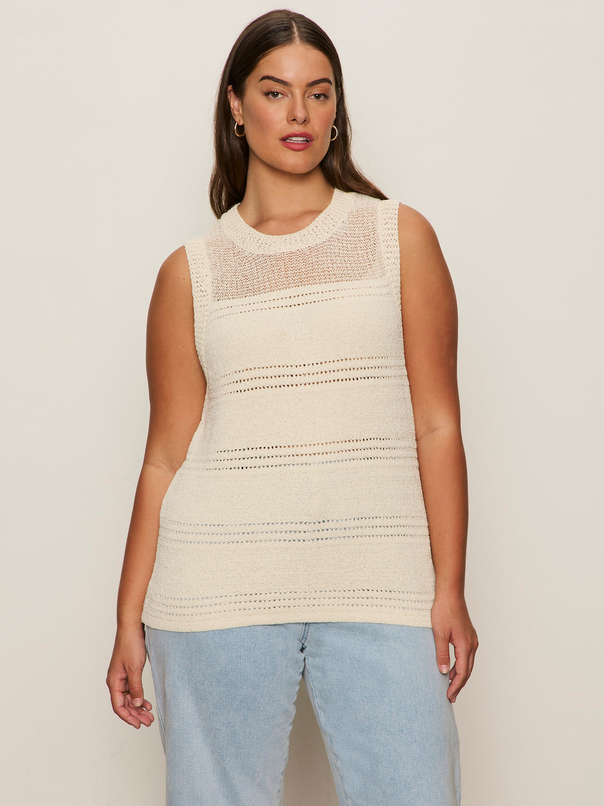 A woman with long brown hair wears the Sanctuary Clothing "waves of summer shell sweater macrame" sleeveless cream knit top and light blue jeans, standing against a plain beige background.