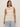 A woman with long brown hair wears the Sanctuary Clothing "waves of summer shell sweater macrame" sleeveless cream knit top and light blue jeans, standing against a plain beige background.